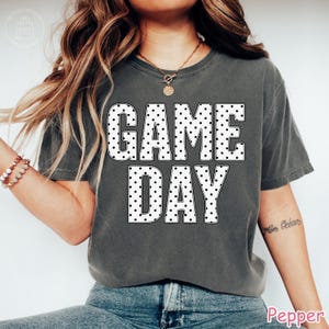 Puede incluir: Una camiseta de manga corta gris carb&oacute;n con el texto "GAME DAY" en letras grandes y blancas con un patr&oacute;n de puntos negros. La prenda tiene cuello redondo, un corte relajado y se combina con pantalones vaqueros azules y un collar dorado.