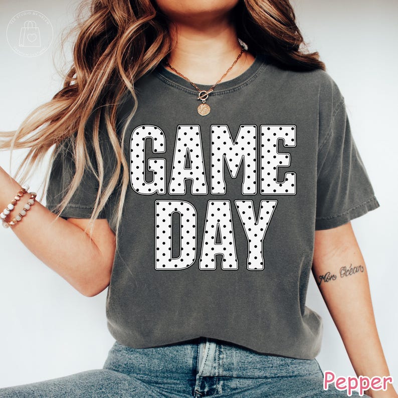 Puede incluir: Una camiseta de manga corta gris carb&oacute;n con el texto "GAME DAY" en letras grandes y blancas con un patr&oacute;n de puntos negros. La prenda tiene cuello redondo, un corte relajado y se combina con pantalones vaqueros azules y un collar dorado.