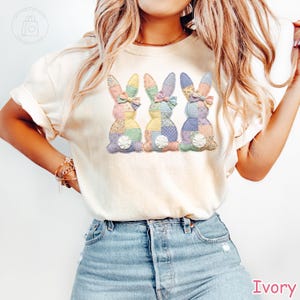 K&ouml;nnte beinhalten: Elfenbeinfarbenes T-Shirt mit drei Patchwork-Hasen mit Pastellmustern und Schleifen. Die Hasen haben wei&szlig;e, flauschige Schw&auml;nze. Das Wort "Ivory" ist unten rechts in Pink gedruckt.