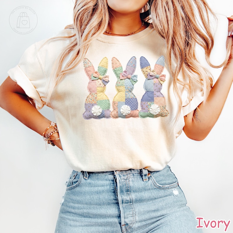 K&ouml;nnte beinhalten: Elfenbeinfarbenes T-Shirt mit drei Patchwork-Hasen mit Pastellmustern und Schleifen. Die Hasen haben wei&szlig;e, flauschige Schw&auml;nze. Das Wort "Ivory" ist unten rechts in Pink gedruckt.