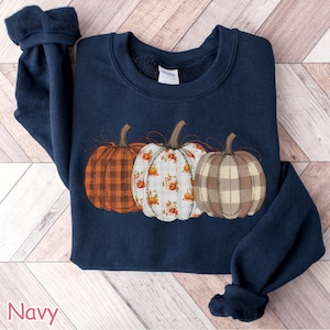 Puede incluir: Sudadera azul marino con tres calabazas decorativas con diferentes dise&ntilde;os: cuadros, floral y cosido. Las calabazas son en tonos naranja, blanco y marr&oacute;n. La palabra "Navy" est&aacute; impresa en la esquina inferior izquierda.