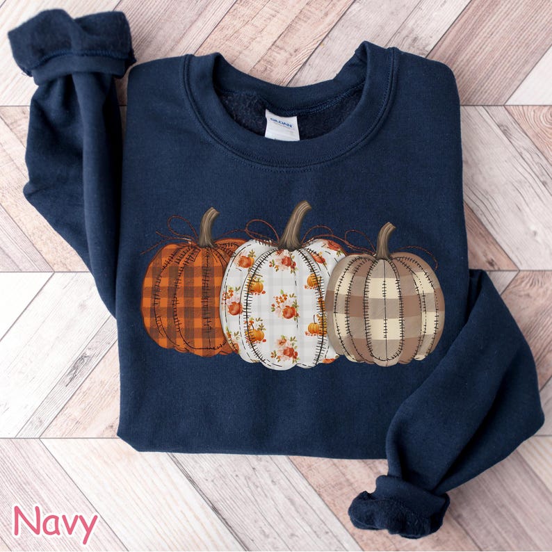 Puede incluir: Sudadera azul marino con tres calabazas decorativas con diferentes dise&ntilde;os: cuadros, floral y cosido. Las calabazas son en tonos naranja, blanco y marr&oacute;n. La palabra "Navy" est&aacute; impresa en la esquina inferior izquierda.