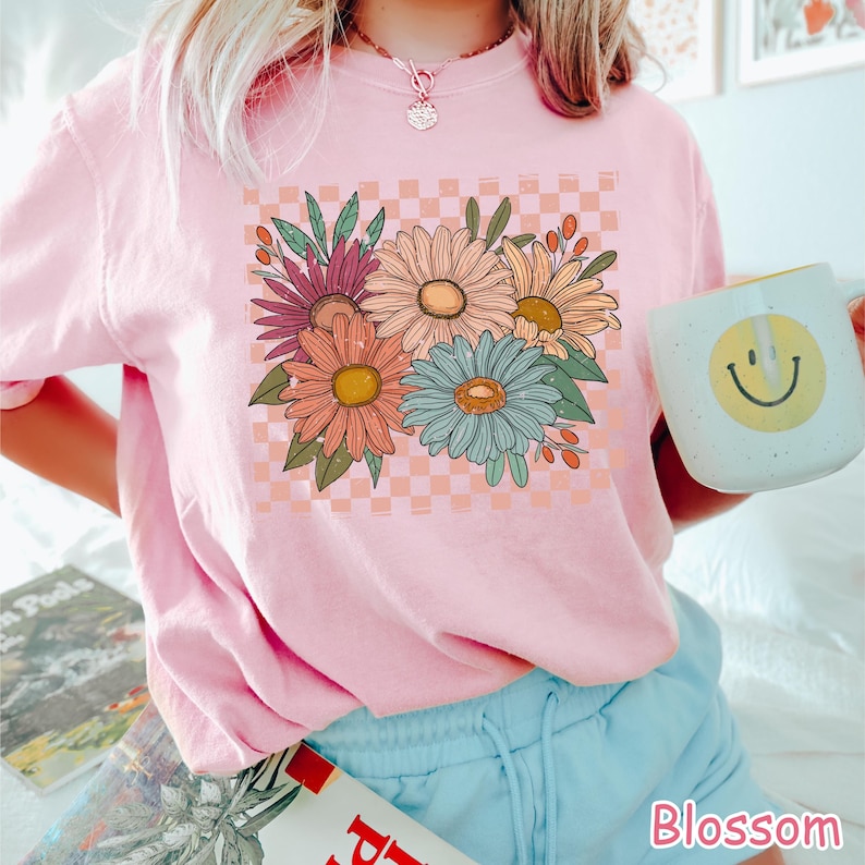 May include: A light pink t-shirt featuring a floral design with daisies in shades of orange, pink, and blue, set against a peach and white checkered background. The word "Blossom" is printed in pink at the bottom.