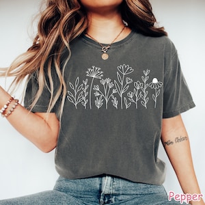 May include: A gray t-shirt with a white outline of a row of flowers and leaves. The shirt is worn by a person with long brown hair.