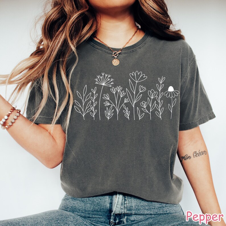 May include: A gray t-shirt with a white outline of a row of flowers and leaves. The shirt is worn by a person with long brown hair.