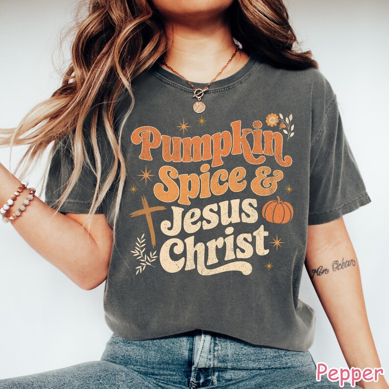 Op de afbeelding: Een donkergrijs t-shirt met de tekst "Pumpkin Spice & Jesus Christ" in oranje en cr&egrave;me. Het ontwerp bevat een kruis, een pompoen en bloemmotieven. Het shirt heeft een casual stijl.