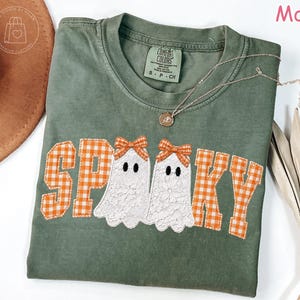 May include: A moss green short-sleeve t-shirt featuring the word SPOOKY in orange and white gingham block letters. Two white lace ghosts with orange gingham bows on their heads replace the letter O, centered on the chest of the garment.