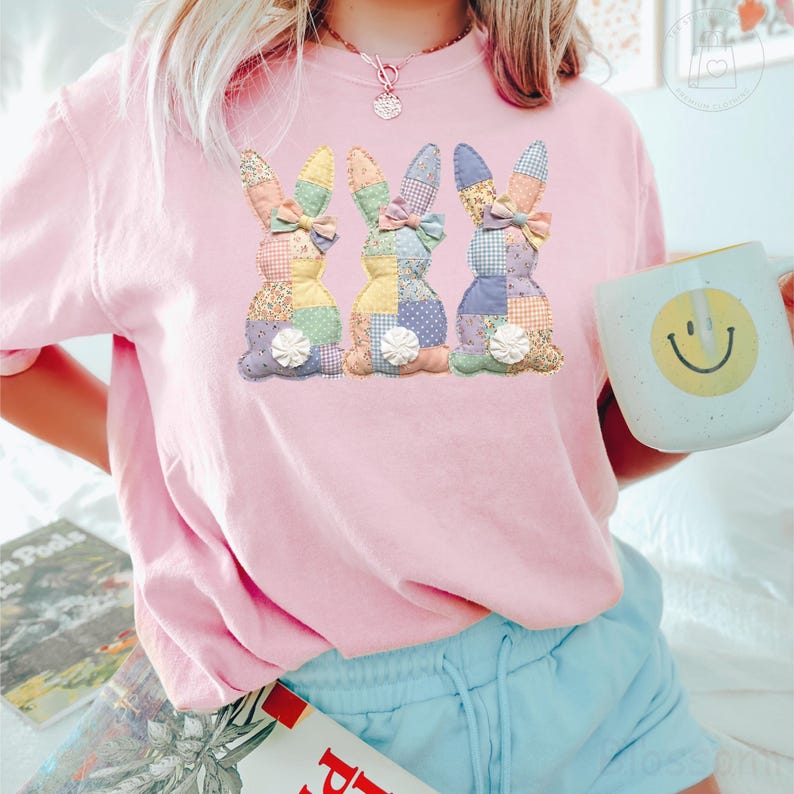 K&ouml;nnte beinhalten: Hellrosa Sweatshirt mit drei Patchwork-Hasen-Designs in Pastellfarben. Jeder Hase hat eine Schleife und eine wei&szlig;e Blume. Eine wei&szlig;e Tasse mit einem gelben Smiley wird in der Hand gehalten.