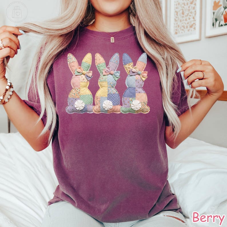 K&ouml;nnte beinhalten: Beerenfarbenes T-Shirt mit drei Patchwork-Hasen-Designs mit bunten Ohren und Schleifen. Die Hasen haben wei&szlig;e, flauschige Schw&auml;nze. Das Shirt ist ein bequemer, l&auml;ssiger Stil.