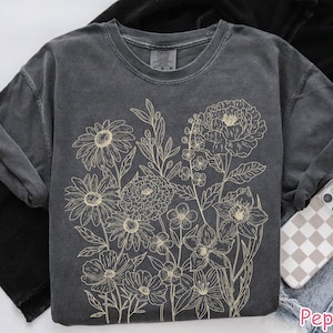 Puede incluir: Una camiseta gris oscuro con un dise&ntilde;o floral beige. El dise&ntilde;o incluye varias flores y hojas. La camiseta est&aacute; dise&ntilde;ada con mangas enrolladas y se combina con una funda de tel&eacute;fono con un patr&oacute;n de cuadros.