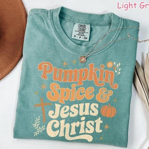 Op de afbeelding: Lichtgroen t-shirt met de woorden "Pumpkin Spice & Jesus Christ" in oranje en cr&egrave;mekleurige letters. Het shirt heeft een kruis, een pompoen en bloemmotieven. Een zilveren ketting met een hanger is over het shirt gedrapeerd.