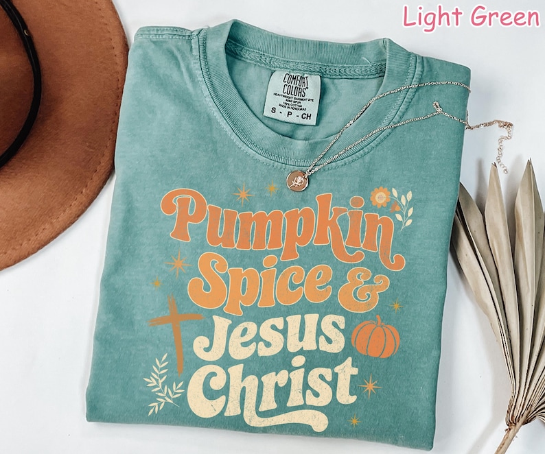 Op de afbeelding: Lichtgroen t-shirt met de woorden "Pumpkin Spice & Jesus Christ" in oranje en cr&egrave;mekleurige letters. Het shirt heeft een kruis, een pompoen en bloemmotieven. Een zilveren ketting met een hanger is over het shirt gedrapeerd.