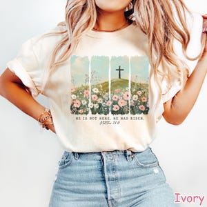 May include: A white t-shirt with a floral design and a cross. The text "He is not here, He has risen. Matthew 28:6" is printed on the shirt.