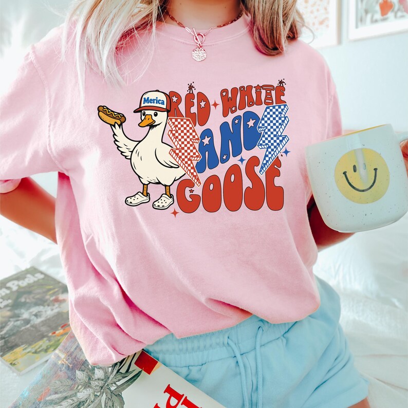 May include: Pink t-shirt with a graphic of a goose wearing a hat that says "Merica" and holding a hot dog. The text "Red, White, and Goose" is in red, white, and blue with lightning bolt graphics. A white mug with a yellow smiley face is also visible.