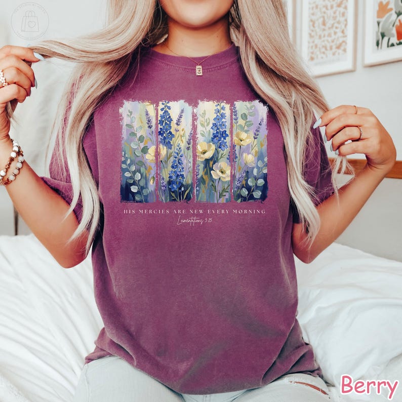 K&ouml;nnte beinhalten: Beerenfarbenes T-Shirt mit einem Blumendesign mit blauen, gelben und wei&szlig;en Blumen. Das Design ist in vertikale Paneele unterteilt. Der Text "HIS MERCIES ARE NEW EVERY MORNING Lamentations 3:23" ist darunter gedruckt.