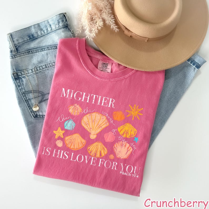 K&ouml;nnte beinhalten: Ein pinkfarbenes T-Shirt mit dem Text "MIGHTIER than the waves of the sea IS HIS LOVE FOR YOU" und Muschelgrafiken. Das Shirt wird mit hellblauen Jeans und einem beigefarbenen Hut kombiniert.