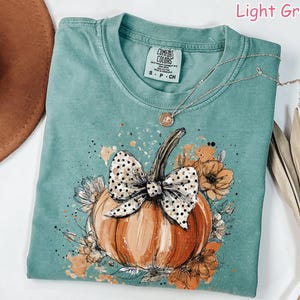 May include: Light green Comfort Colors t-shirt with a pumpkin graphic. The pumpkin is orange with a black and white polka dot bow. The shirt has a crew neck and short sleeves. A silver necklace with a pendant is on the shirt.