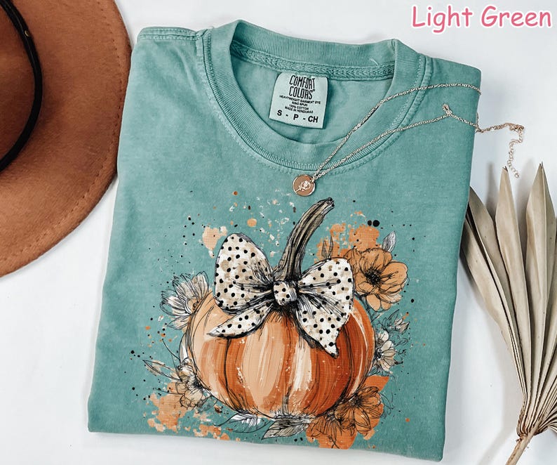 May include: Light green Comfort Colors t-shirt with a pumpkin graphic. The pumpkin is orange with a black and white polka dot bow. The shirt has a crew neck and short sleeves. A silver necklace with a pendant is on the shirt.