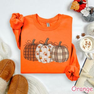 Puede incluir: Una sudadera naranja con un dise&ntilde;o de tres calabazas decorativas con patrones de cuadros y flores. Las calabazas tienen tallos marrones y est&aacute;n cosidas juntas. La palabra "Orange" est&aacute; escrita en rosa en la parte inferior derecha.