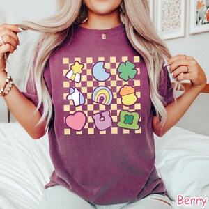 Peut inclure: Un t-shirt violet &agrave; motif &agrave; carreaux avec une vari&eacute;t&eacute; d'images de dessins anim&eacute;s color&eacute;s, notamment un arc-en-ciel, un fer &agrave; cheval, un tr&egrave;fle &agrave; quatre feuilles, un pot d'or, une licorne et une &eacute;toile.