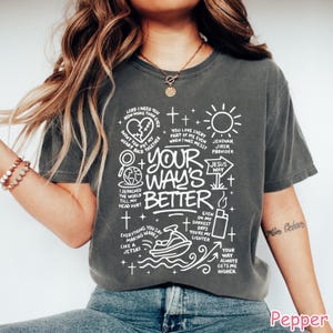 Op de afbeelding: Donkergrijs t-shirt met witte grafische tekst en illustraties. Het ontwerp bevat zinnen als "Your Way's Better", "Jesus Way" en "Even on my darkest days you're my lighter". Het t-shirt heeft een relaxte pasvorm.