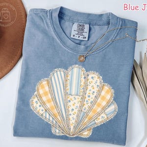 May include: Blue jean colored t-shirt with a seashell design. The seashell is made of various patterned fabrics, including stripes, gingham, and floral prints. A gold necklace with a pendant rests on the shirt.