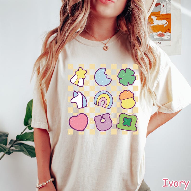 Peut inclure: Un t-shirt cr&egrave;me avec un motif &agrave; carreaux et un graphique de neuf images de dessins anim&eacute;s color&eacute;s. Les images comprennent un arc-en-ciel, un fer &agrave; cheval, un tr&egrave;fle &agrave; quatre feuilles, un pot d'or, une licorne, un c&oelig;ur, une &eacute;toile, un croissant de lune et un tr&egrave;fle.