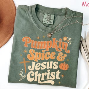 Op de afbeelding: Een mosgroen t-shirt met de woorden "Pumpkin Spice & Jesus Christ" in oranje en cr&egrave;mekleurige letters. Het shirt heeft een kruis en een pompoenafbeelding. Een gouden ketting is over het shirt gedrapeerd.