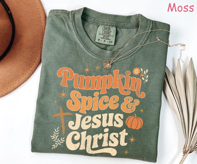 Op de afbeelding: Een mosgroen t-shirt met de woorden "Pumpkin Spice & Jesus Christ" in oranje en cr&egrave;mekleurige letters. Het shirt heeft een kruis en een pompoenafbeelding. Een gouden ketting is over het shirt gedrapeerd.