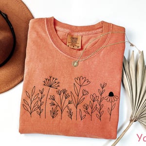 May include: A burnt orange t-shirt with a black line drawing of wildflowers on the front. The shirt has a crew neck and is made of a soft, comfortable fabric. The text "Comfort Colors" is visible on the tag.