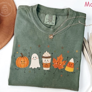 Op de afbeelding: Een mosgroen t-shirt met korte mouwen en een grafische print van vijf lachende herfstfiguren: een oranje pompoen, een wit spookje, een koffiebeker met slagroom, een oranje esdoornblad en een stukje maissnoep.