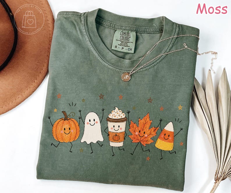 Op de afbeelding: Een mosgroen t-shirt met korte mouwen en een grafische print van vijf lachende herfstfiguren: een oranje pompoen, een wit spookje, een koffiebeker met slagroom, een oranje esdoornblad en een stukje maissnoep.