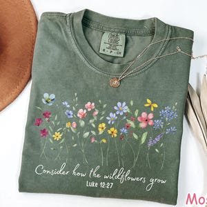 May include: A moss green t-shirt with a floral design and the text "Consider how the wildflowers grow, Luke 12:27." The shirt has a crew neck and short sleeves. A gold necklace with a pendant is on the shirt.