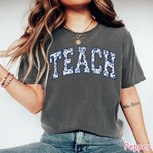 May include: A dark gray t-shirt with the word "TEACH" printed in white with a blue floral pattern. The shirt has a relaxed fit and short sleeves.