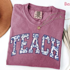 May include: A maroon t-shirt with the word "TEACH" in white letters outlined in navy blue. The letters are filled with a blue and white floral pattern. The shirt is made by Comfort Colors and is size S-P-CH.