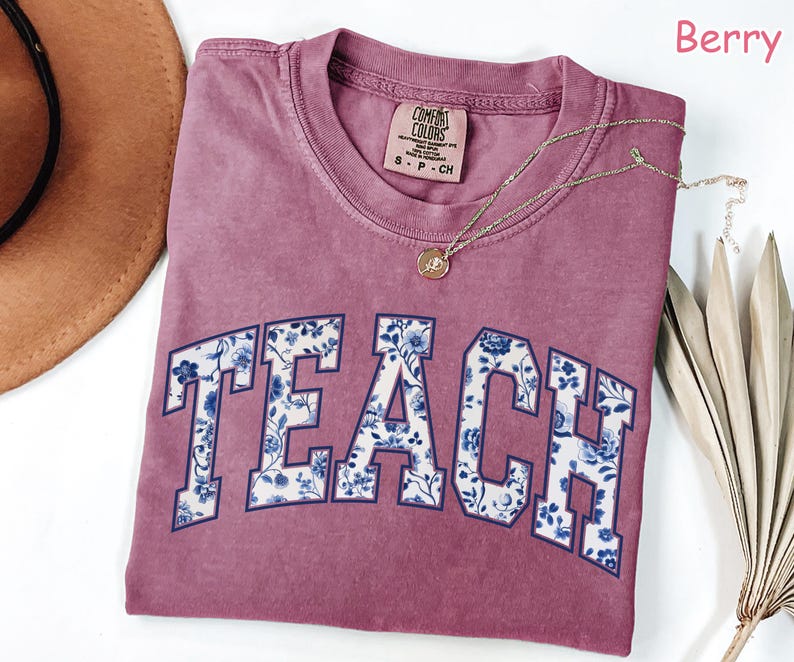 May include: A maroon t-shirt with the word "TEACH" in white letters outlined in navy blue. The letters are filled with a blue and white floral pattern. The shirt is made by Comfort Colors and is size S-P-CH.