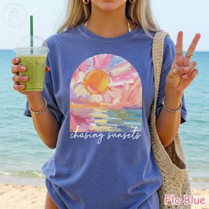 May include: A blue t-shirt with a colorful sunset graphic inside an arch. The graphic features a yellow sun, pink and orange clouds, and the text "chasing sunsets" in white script. The shirt is labeled "Flo Blue".