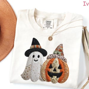 May include: Ivory-colored t-shirt featuring a Halloween-themed design. The design includes a ghost wearing a witch hat and a jack-o'-lantern. The shirt has a crew neck and short sleeves. The brand label is visible on the shirt.
