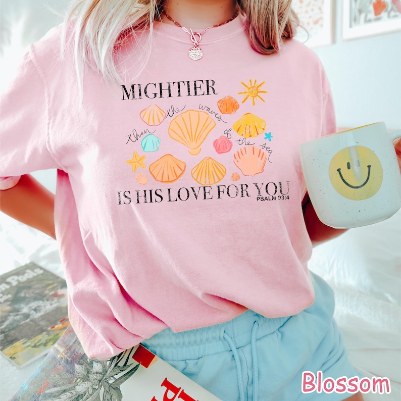 K&ouml;nnte beinhalten: Hellrosa Sweatshirt mit dem Text "MIGHTIER than the waves of the sea IS HIS LOVE FOR YOU" und Muschel-Grafiken. Eine wei&szlig;e Tasse mit einem gelben Smiley wird gehalten. Das Wort "Blossom" befindet sich in der unteren rechten Ecke.