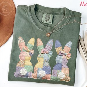 K&ouml;nnte beinhalten: Ein moosgr&uuml;nes T-Shirt mit drei aufgen&auml;hten Hasen, jeweils mit Patchwork-Designs und Schleifen. Die Hasen haben bunte Ohren und K&ouml;rper. Eine goldene Halskette mit Anh&auml;nger liegt auf dem Shirt. Das Shirt ist von der Marke Comfort Colors.