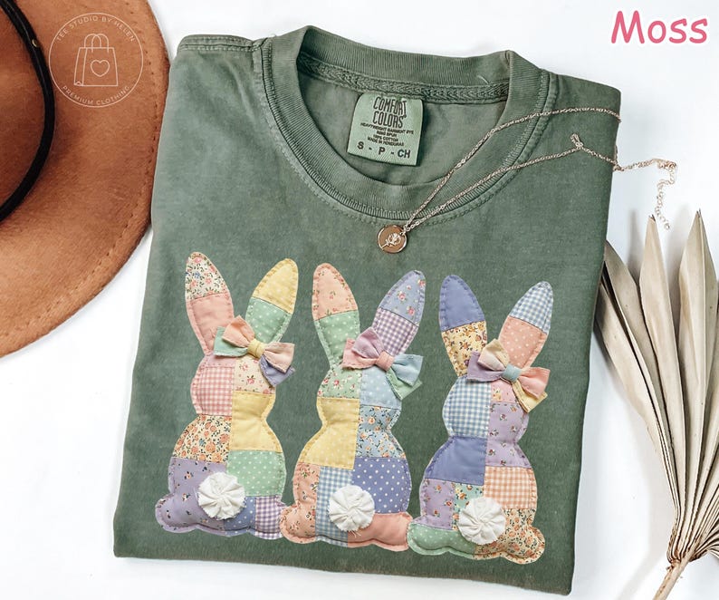 K&ouml;nnte beinhalten: Ein moosgr&uuml;nes T-Shirt mit drei aufgen&auml;hten Hasen, jeweils mit Patchwork-Designs und Schleifen. Die Hasen haben bunte Ohren und K&ouml;rper. Eine goldene Halskette mit Anh&auml;nger liegt auf dem Shirt. Das Shirt ist von der Marke Comfort Colors.