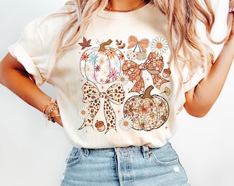 May include: Ivory-colored t-shirt featuring a fall-themed graphic. The design includes floral pumpkins, bows, and daisies in shades of orange, brown, and pink. The word "Ivory" is printed at the bottom right.