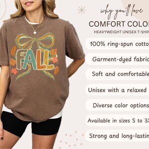 May include: Brown heavyweight unisex t-shirt with the word "FALL" in teal, orange, and brown, surrounded by a bow and autumn leaves. The shirt is made of 100% ring-spun cotton and is available in sizes S to 3XL.