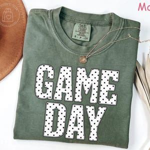 Puede incluir: Una camiseta de cuello redondo color verde musgo con el texto GAME DAY en letras grandes blancas con un patr&oacute;n de puntos negros. La prenda tiene una etiqueta Comfort Colors en el cuello interior y se muestra con un collar dorado y un sombrero marr&oacute;n.