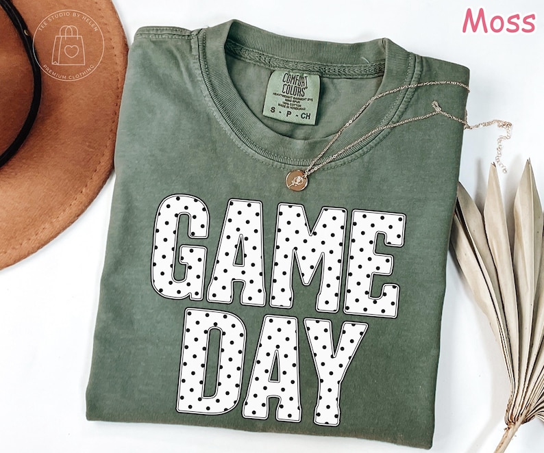 Puede incluir: Una camiseta de cuello redondo color verde musgo con el texto GAME DAY en letras grandes blancas con un patr&oacute;n de puntos negros. La prenda tiene una etiqueta Comfort Colors en el cuello interior y se muestra con un collar dorado y un sombrero marr&oacute;n.