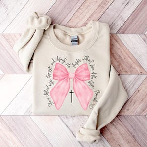 May include: A beige sweatshirt with a pink bow and a cross. The text "She is clothed with strength and dignity; she laughs without fear of the future." is printed around the bow.