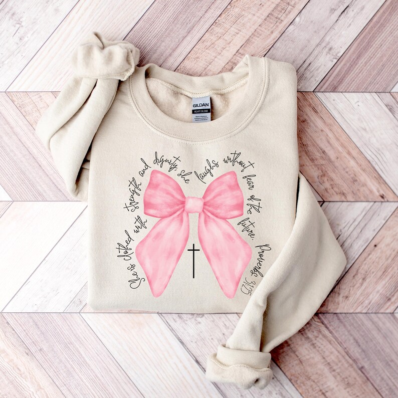 May include: A beige sweatshirt with a pink bow and a cross. The text "She is clothed with strength and dignity; she laughs without fear of the future." is printed around the bow.