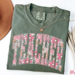 Puede incluir: Una camiseta verde con un estampado floral que deletrea "Teacher" en una fuente retro. La camiseta est&aacute; hecha de un material suave y c&oacute;modo y tiene un corte holgado.