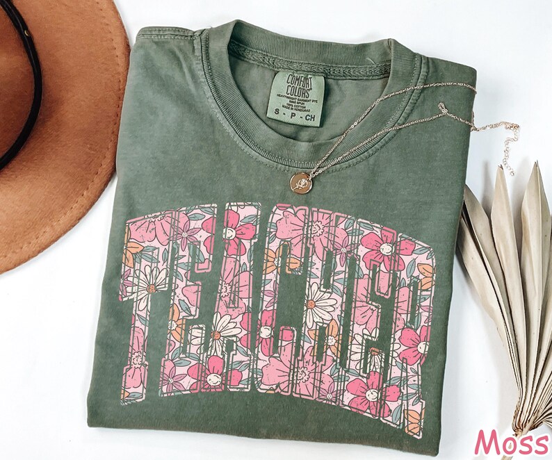 Puede incluir: Una camiseta verde con un estampado floral que deletrea "Teacher" en una fuente retro. La camiseta est&aacute; hecha de un material suave y c&oacute;modo y tiene un corte holgado.