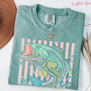 May include: A light green short-sleeved t-shirt featuring a graphic of a fish jumping out of water. The fish is outlined in vibrant colours including yellow, purple, and blue. Behind the fish are vertical pink stripes. The shirt has a crew neckline.